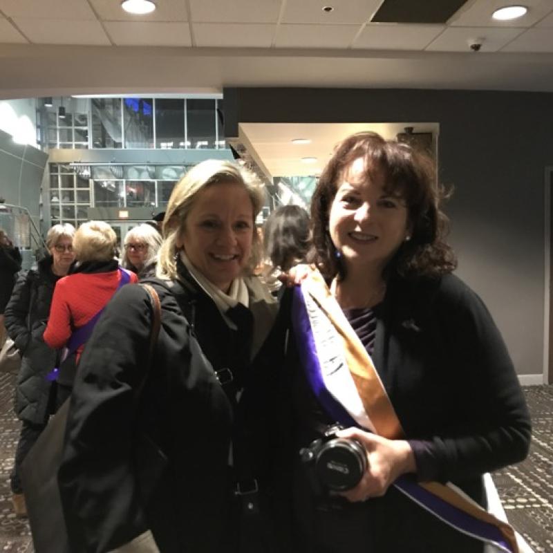 Two women stand indoors smiling.