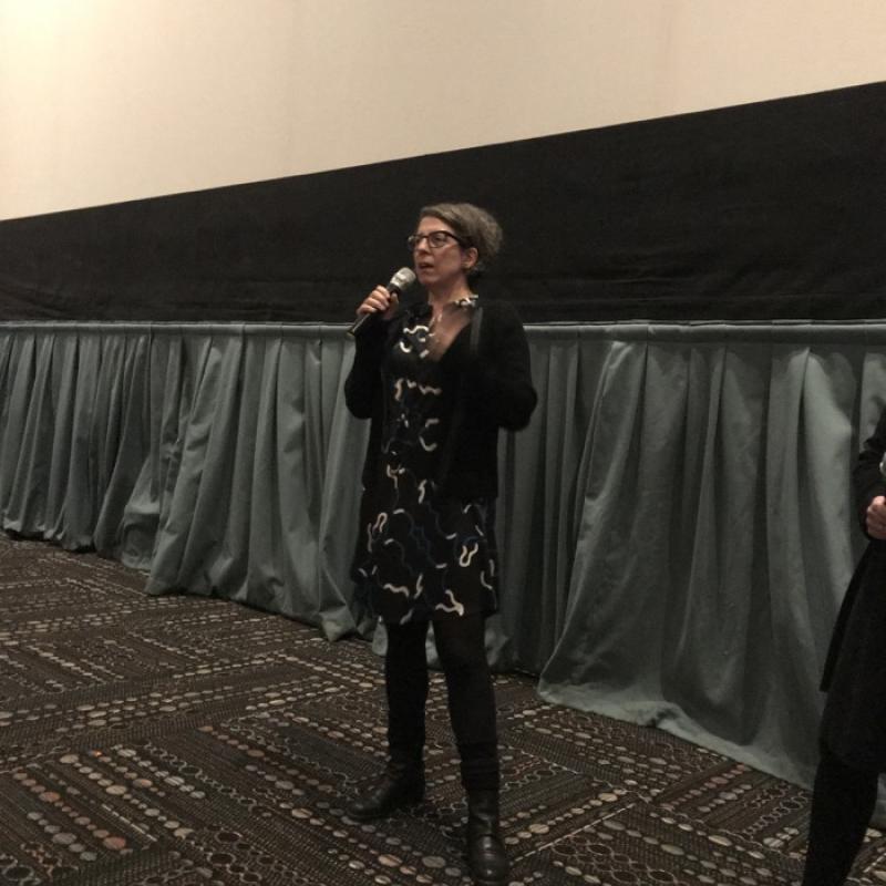 Woman stands in movie theater, speaking in microphone.