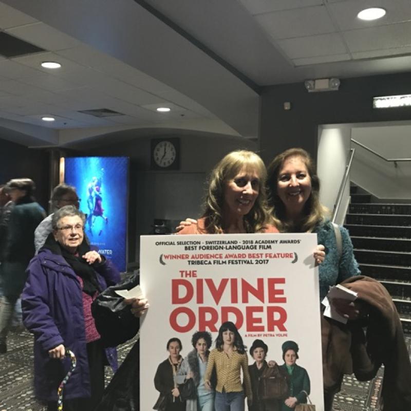 Two women stand indoors smiling next to poster for The Divine Order film.