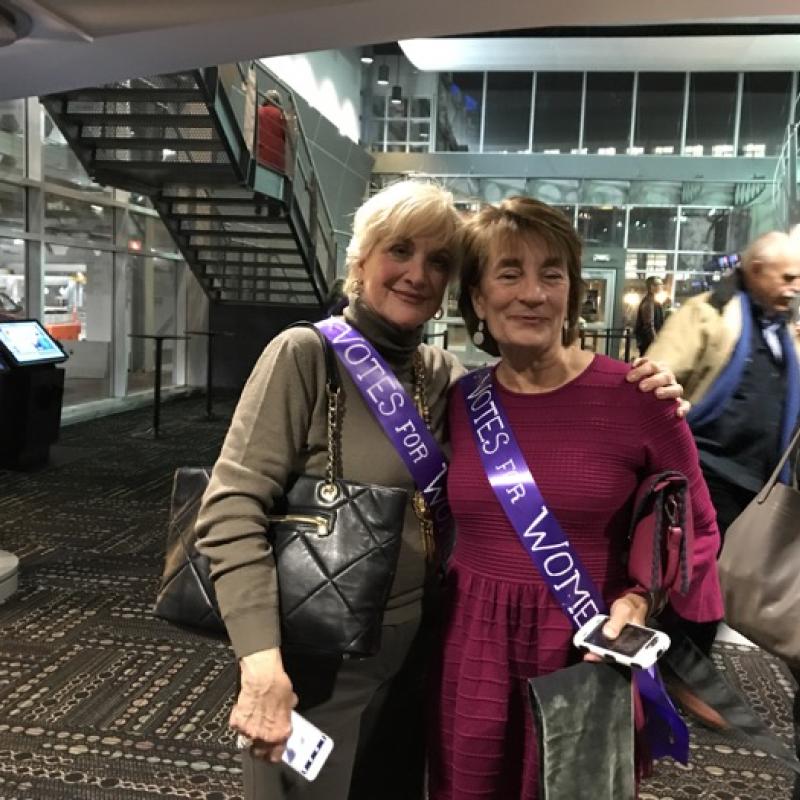 Two women stand indoors smiling.