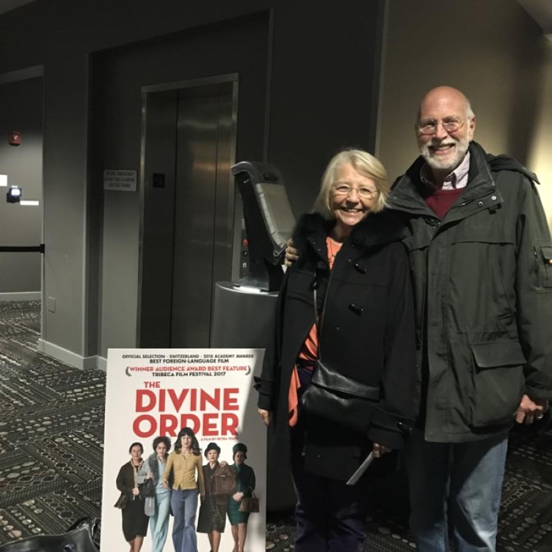 Man and woman stand next to poster for The Divine Order film.