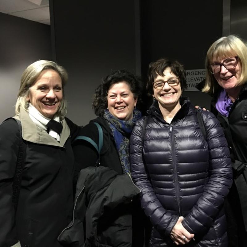 Four women stand indoors smiling.