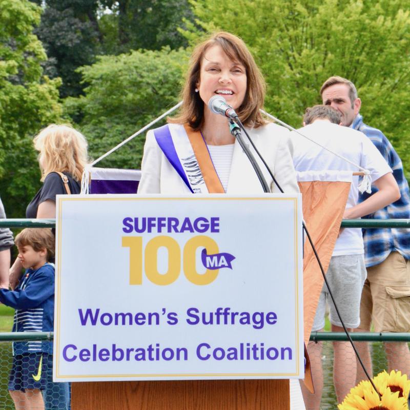 Woman wearing purple sash stands outside speaking at lectern.