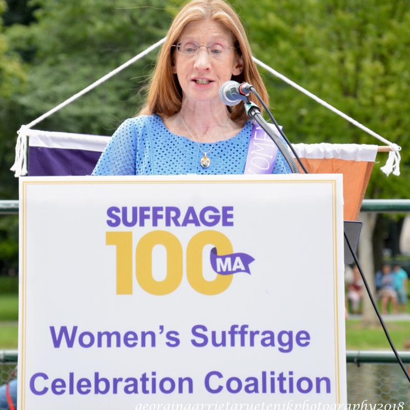 Woman wearing purple sash stands outside speaking at lectern.