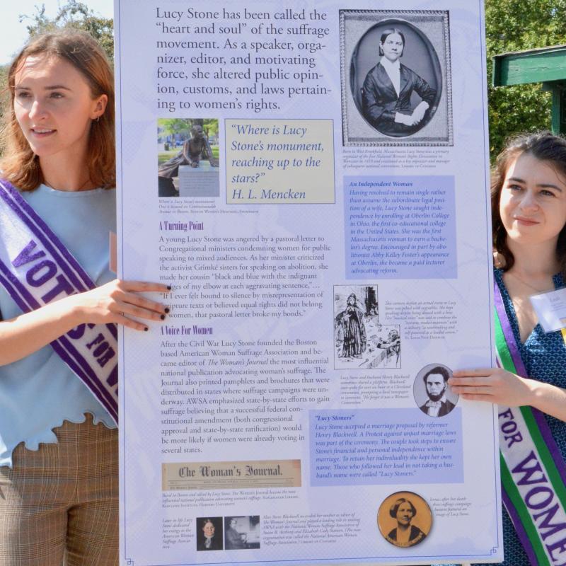 Two women stand next to tall white sign.