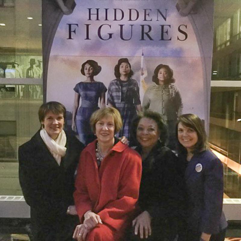 Four women stand in front of Hidden Figures movie poster.