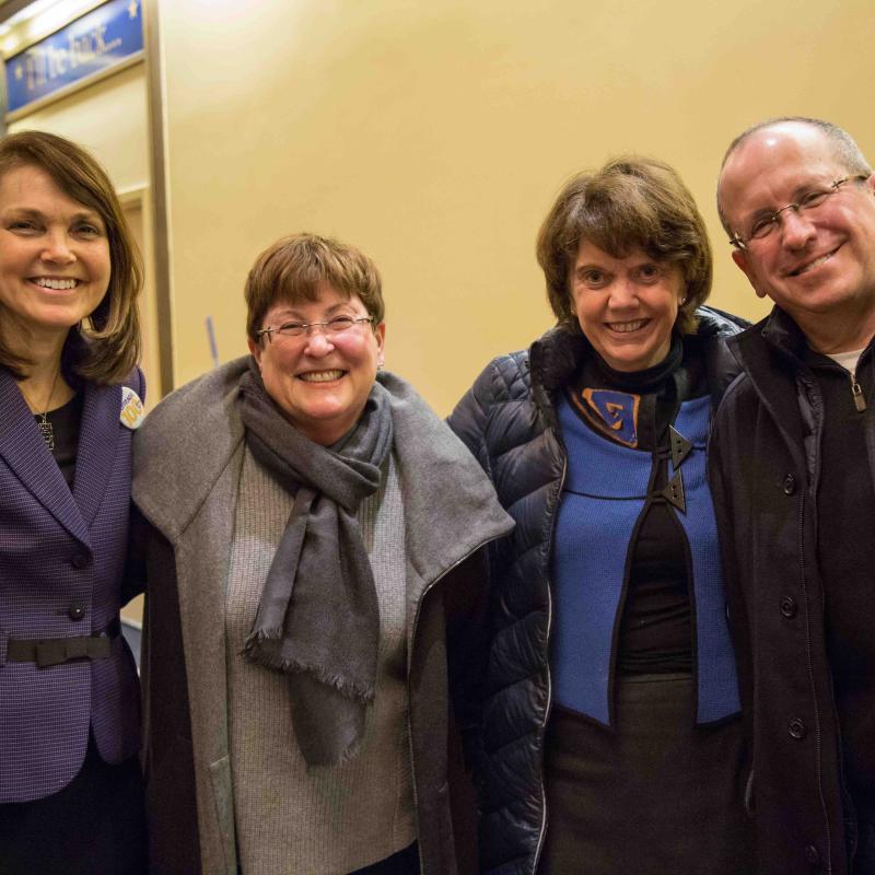 Three women and one man stand indoors smiling.