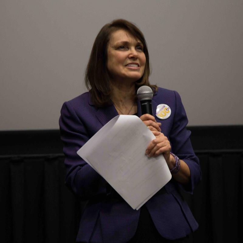 Woman stands in front of screen holding microphone.