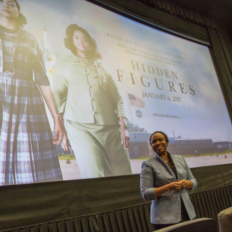 Woman stands in front of screen holding microphone.