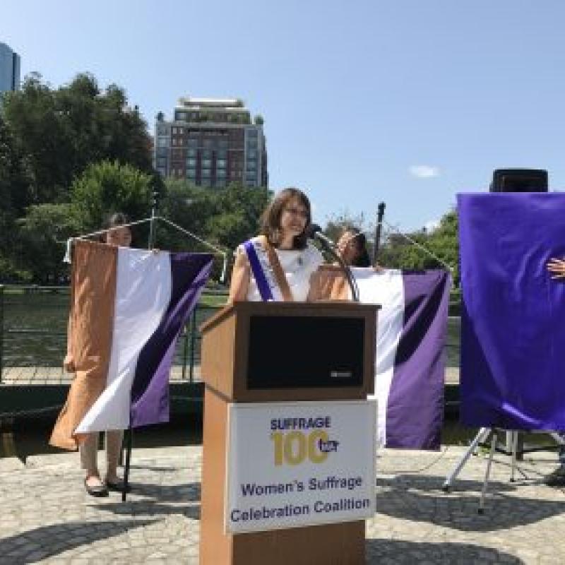 Woman wearing purple sash stands outside speaking at lectern.