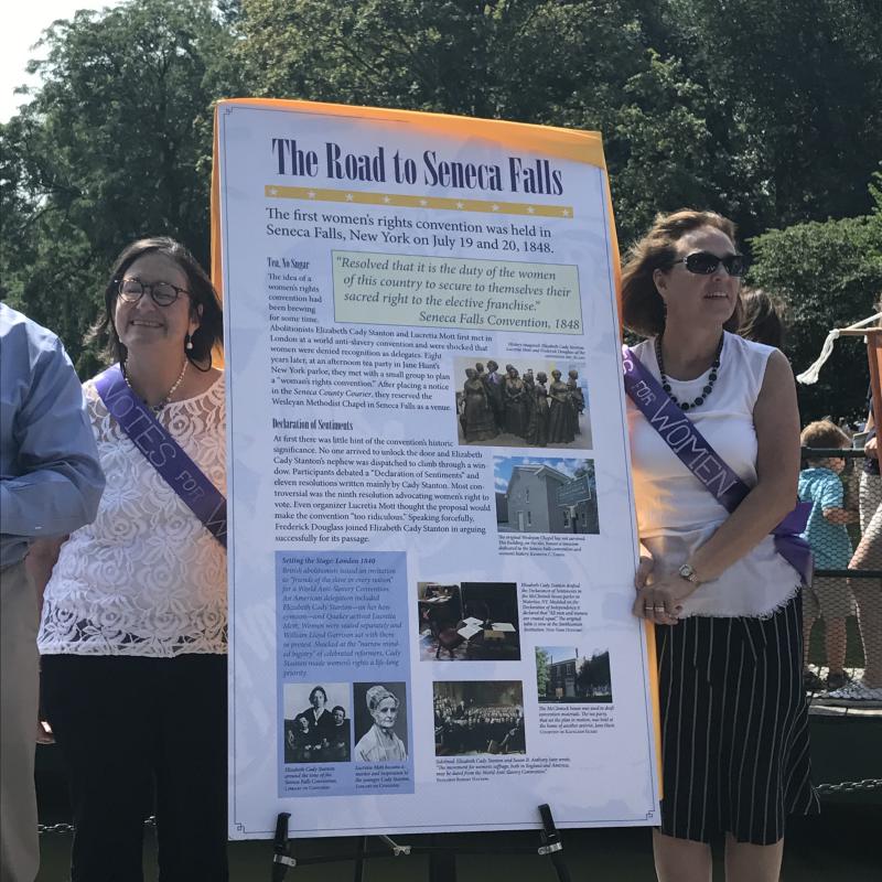 Two women stand next to tall white sign.