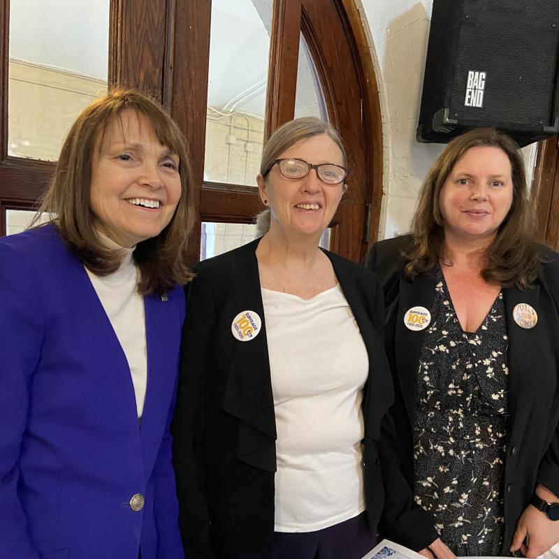 Three women stand indoors smiling.