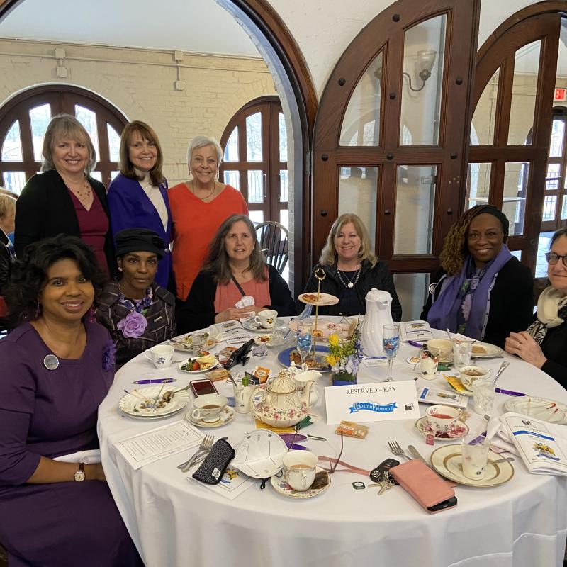 Group of women gather around white table smiling.