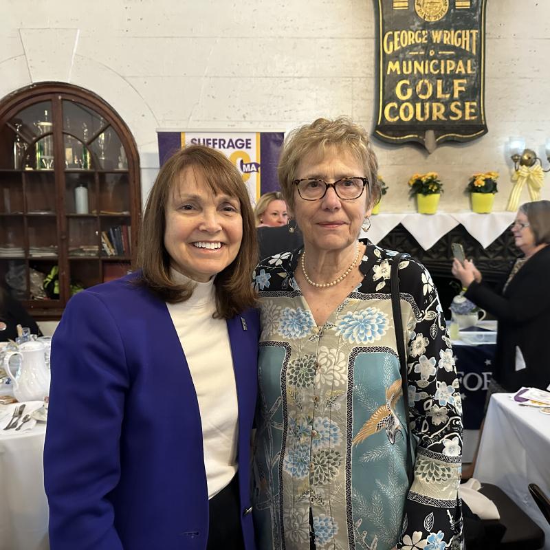 Two women stand indoors smiling.