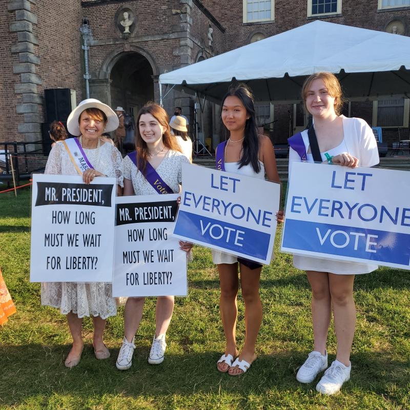 Four young women stand outside holding signs.