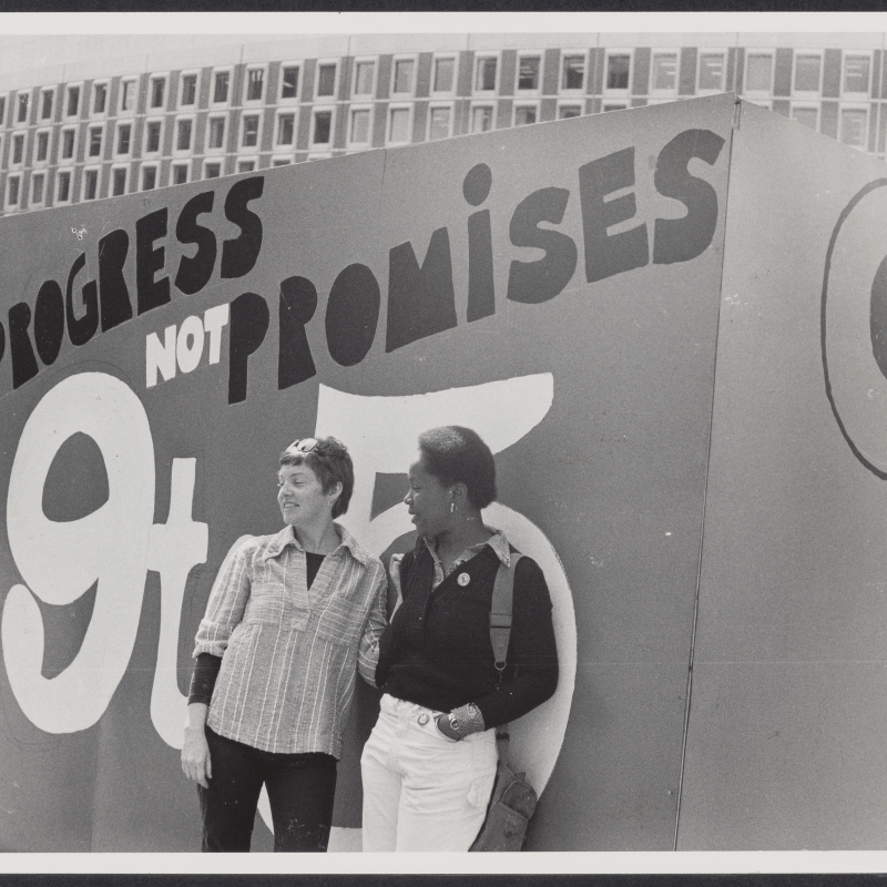 Two women standing outside in front of a massive sign that reads "Progress not Promises: 9to5"