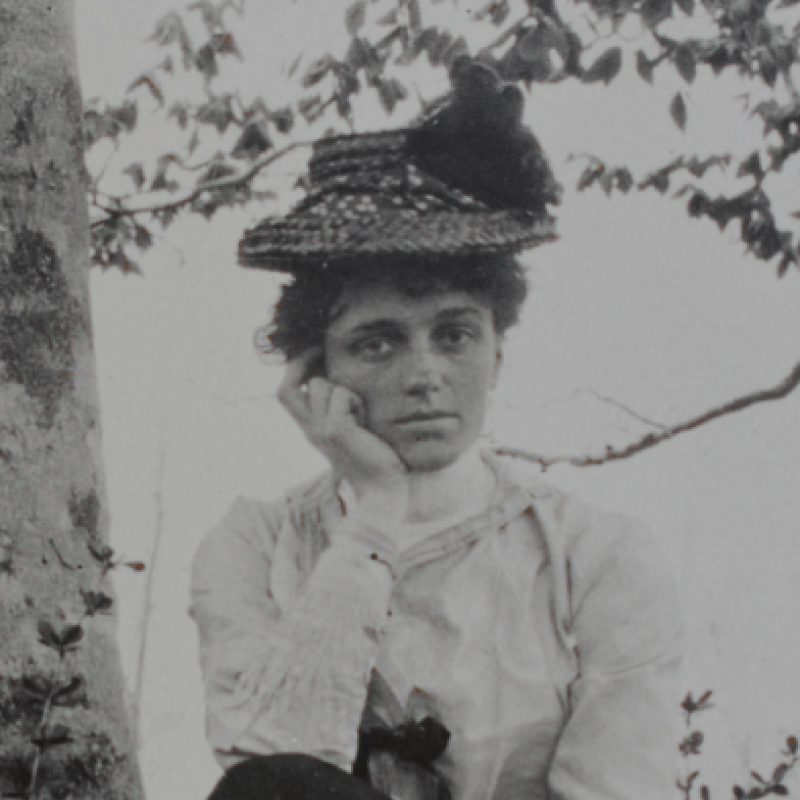 Woman with her hand on her chin, wearing a hat, and sitting on front of a tree.