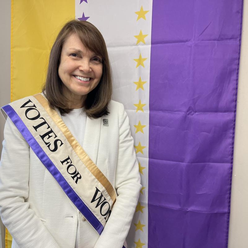 Woman wearing purple sash stands in front of purple and yellow flag.