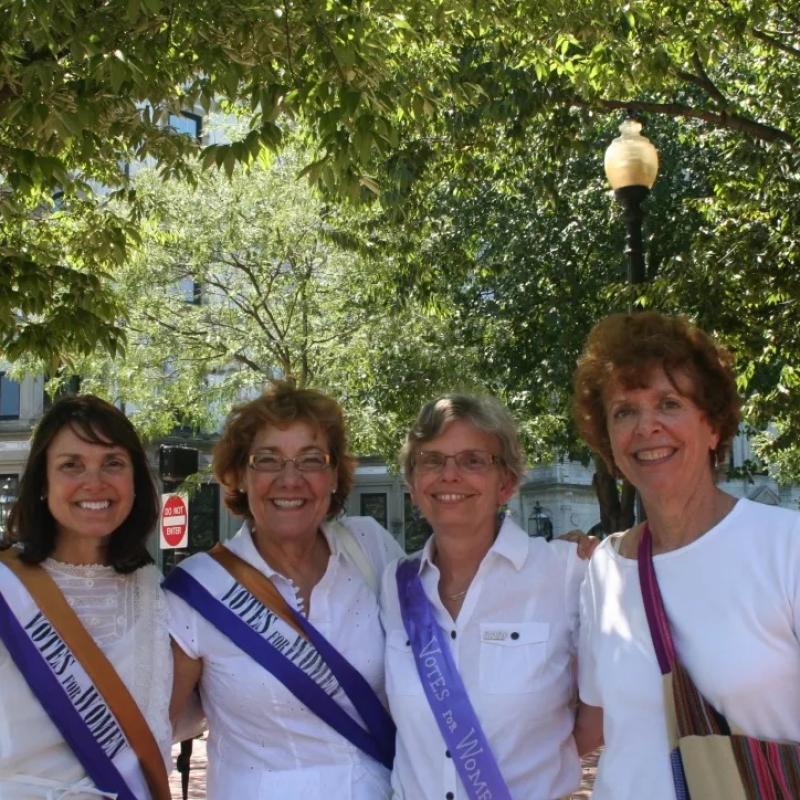 Four women stand outside smiling and wearing sashes.