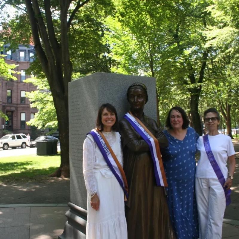 Three women wearing sashes stand next to statue.