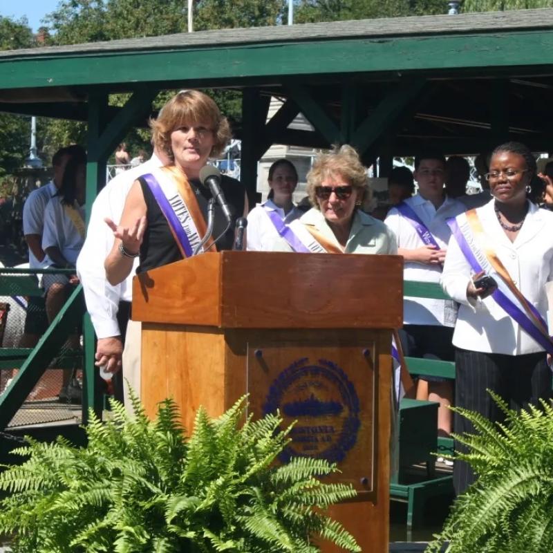 Woman wearing purple sash stands outside speaking at lectern.