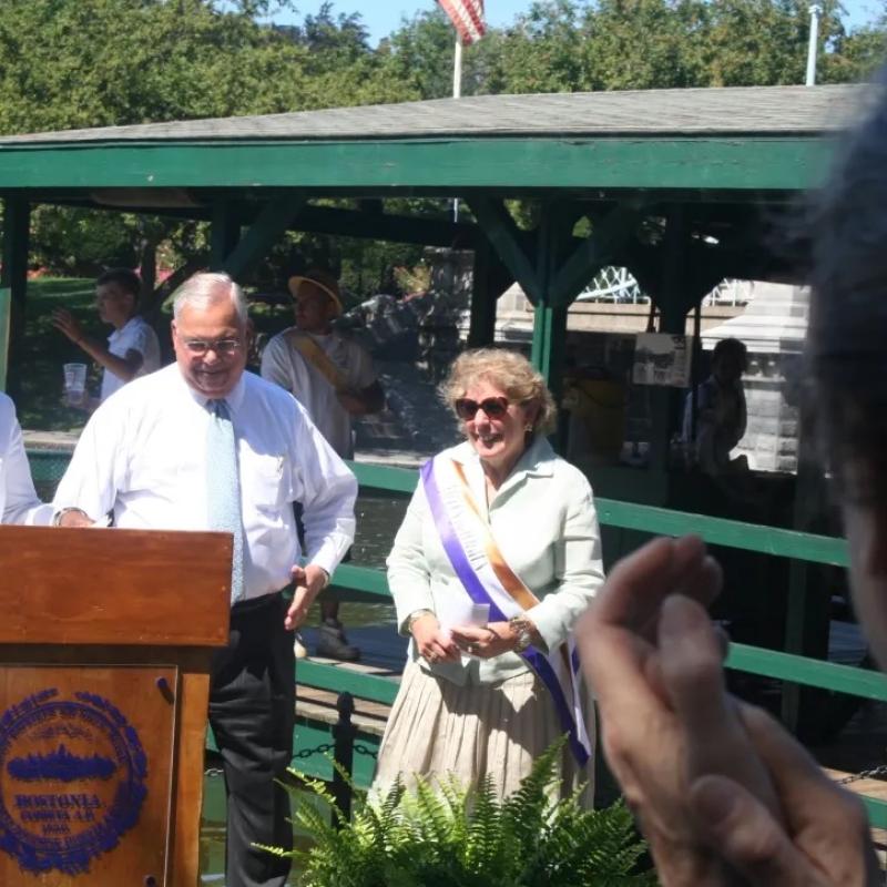 Woman wearing purple sash stands outside speaking at lectern.