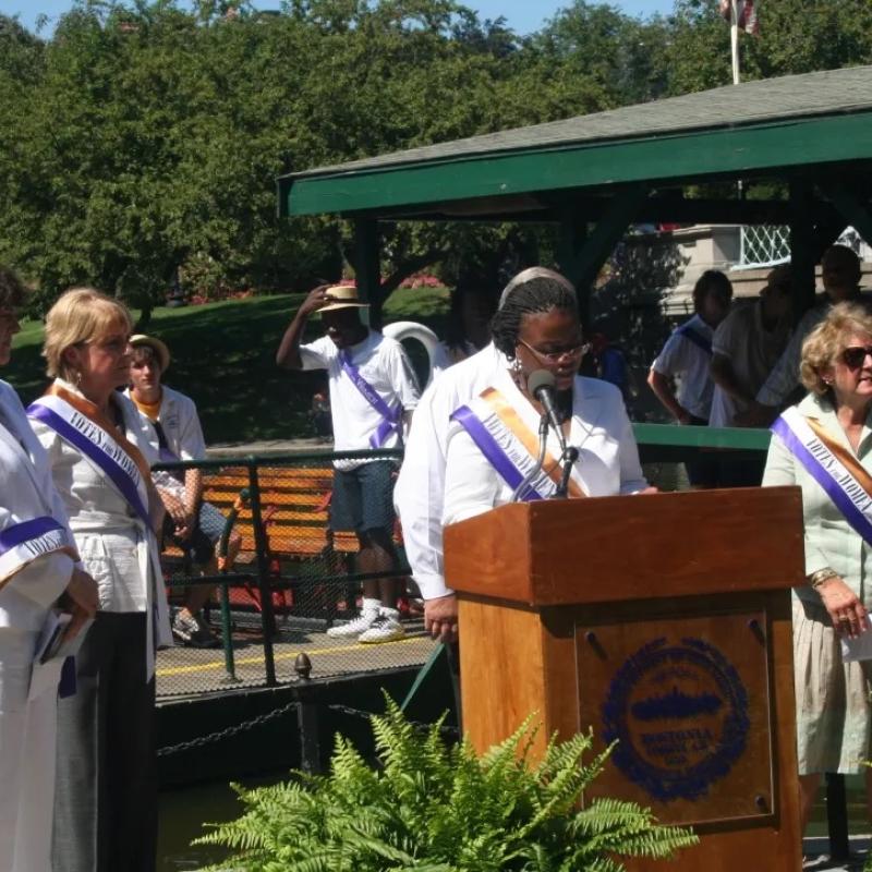 Woman wearing purple sash stands outside speaking at lectern.