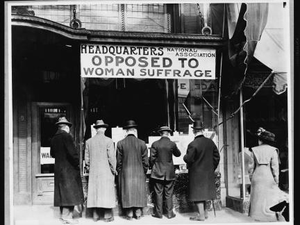 Old photograph of people gathered at table with banner reading, "Headquarters - National Association Opposed to Woman's Suffrage."