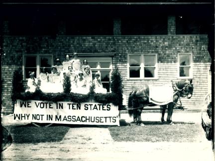 Black and white photo of 10 women in horse-drawn carriage with sign reading, "We vote in ten states. Why not Massachusetts?".