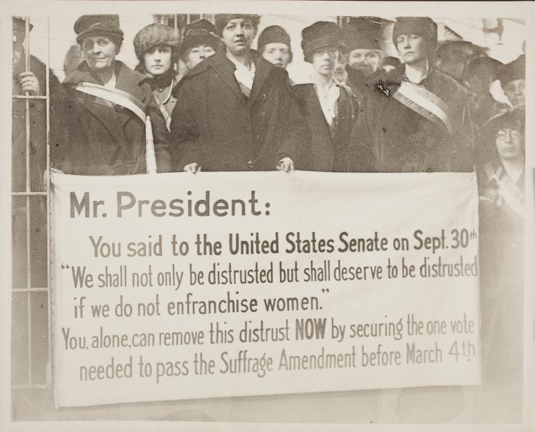 Black and white photo of group of women holding sign addressing President Wilson and women's suffrage.