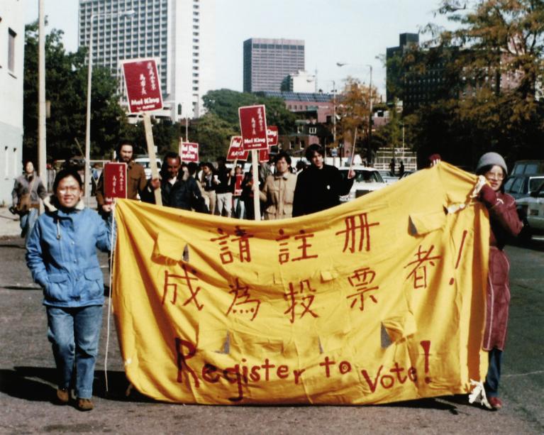 Two women march in Boston holding a yellow banner that reads "Register to Vote" in English and Chinese.