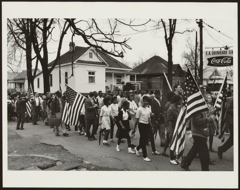 Group of adults in 60s-era clothes marches holding American flags.