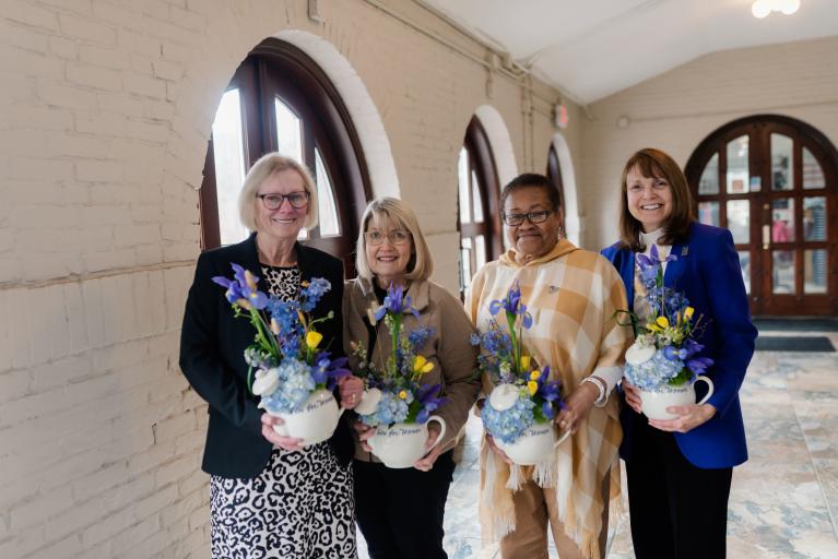 Four women stand indoors smiling and holding flowers.