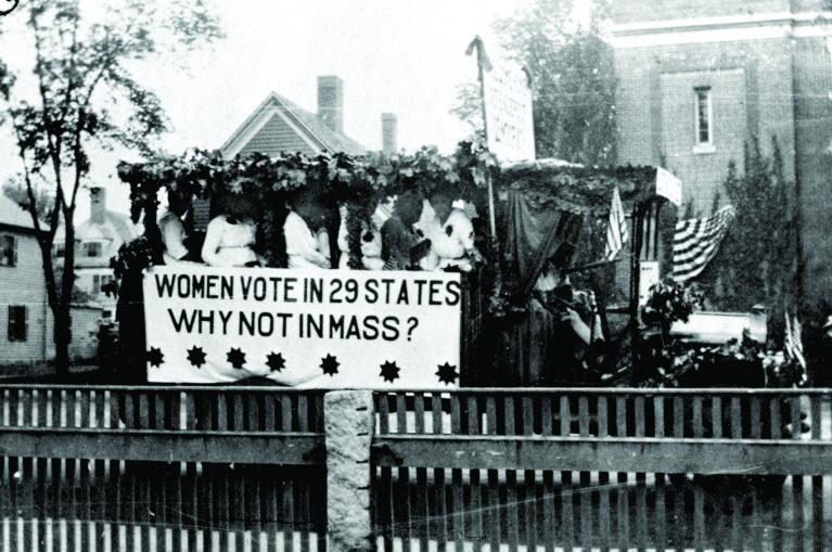 Black and white photo of women on parade float with sign, "Women vote in 29 states. Why not in Mass?".