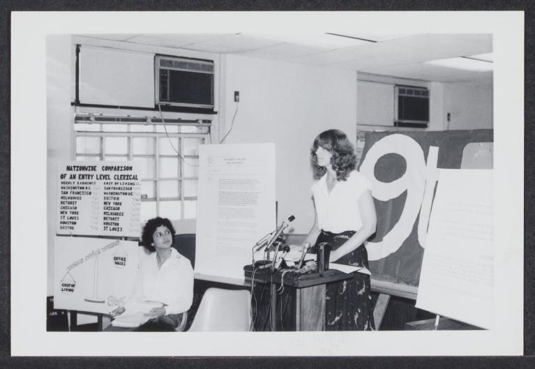 woman with curly hair standing behind a podium in front of a 9to5 sign. Another woman sits off to the side in front of a chart that has earnings and cost of living for different cities.