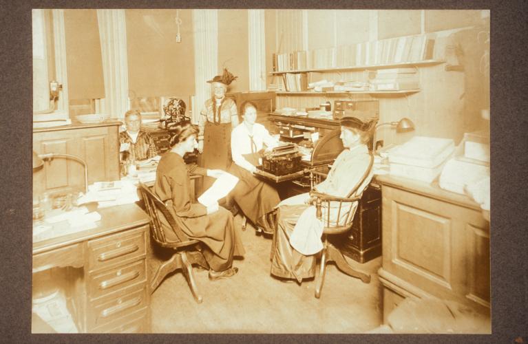 Black and white photograph of Stantial, Luscomb, Blackmur, Needham, and an unidentified woman in the BESAGG office. Three desks are visible in the picture; the office looks a bit cramped.