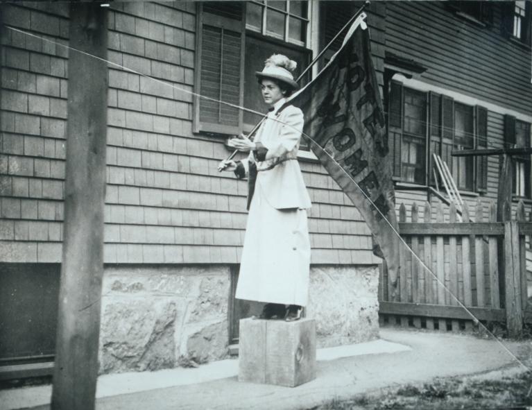 Suffragist Florence Luscomb speaks while standing on a soapbox, holding a votes for women flag.