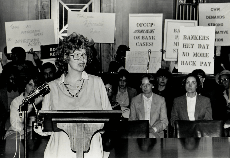 woman with curly hair and glasses stands at a podium giving a speech while a group of women with signs are standing behind her.