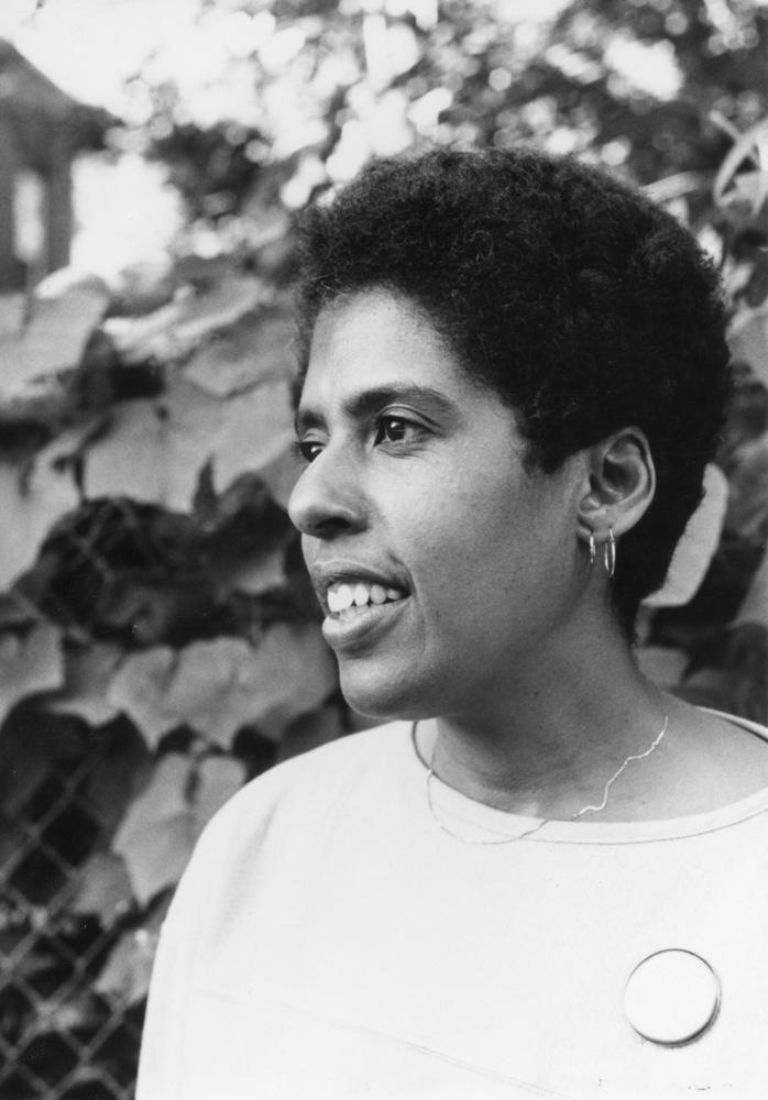 Black and white photograph of Barbara Smith in profile in front of a leaf-covered fence