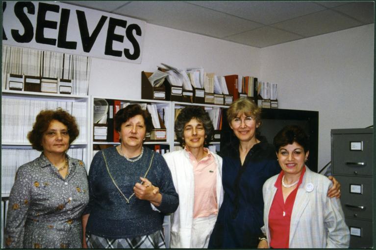 Color photograph of 5 women in front of bookshelves in an office setting.