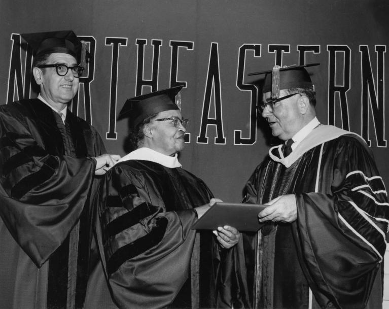 Black and white photo of woman dressed in graduation robes being handed a degree by a man in robes, while another man in robes stands behind.