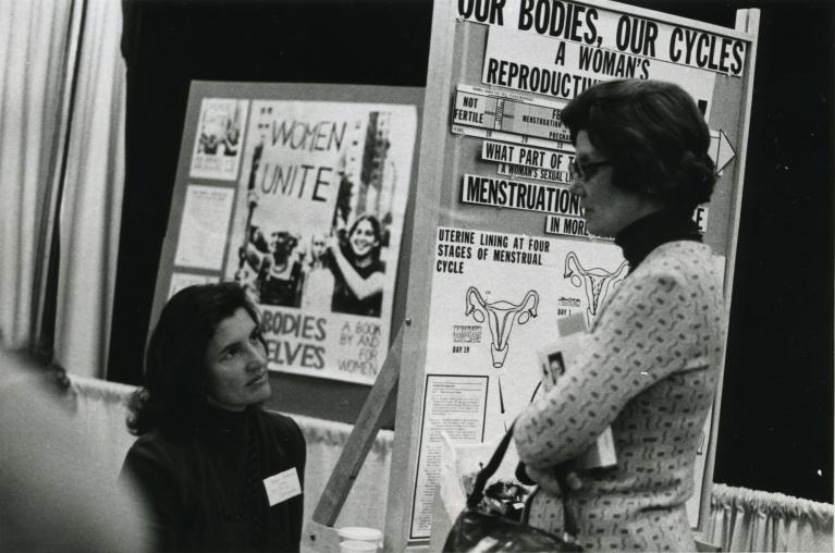 Black and white photograph of two women in front of an exhibit booth for Our Bodies, Ourselves.