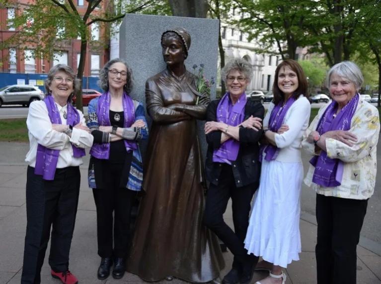 Five women stand outside in front of statue wearing purple scarves.