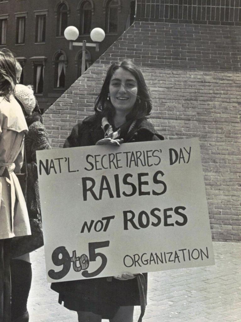 Woman with shoulder-length hair holds a sign that says "Raises not Roses"