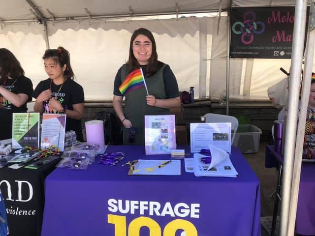 Teen stands at table holding rainbow flag.