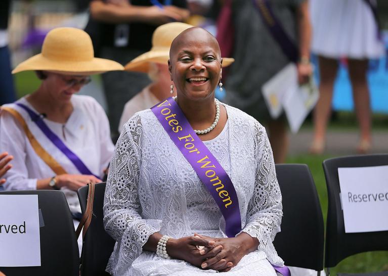 Woman wearing purple sash sits outside smiling.