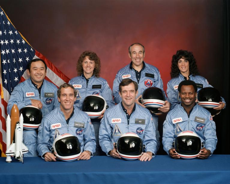 Seven adults in NASA flight suits holding helmets smile in front of red backdrop.
