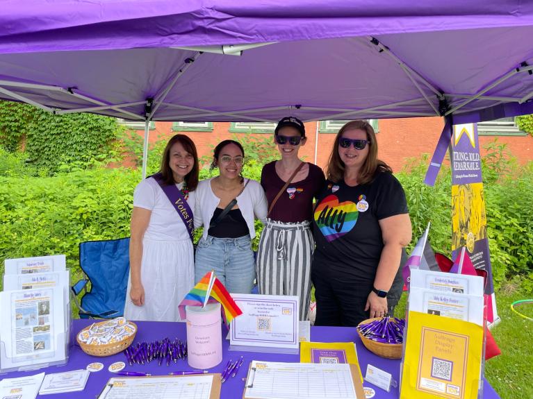 Four women stand at purple table.