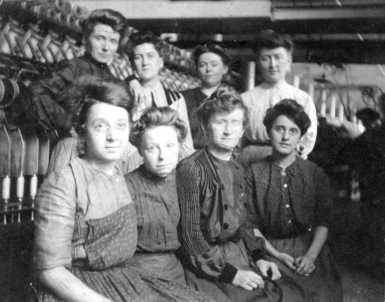 group of 8 women mill workers, four seated and four standing, in a factory