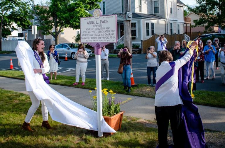 Two women wearing purple sashes stand outside smiling.